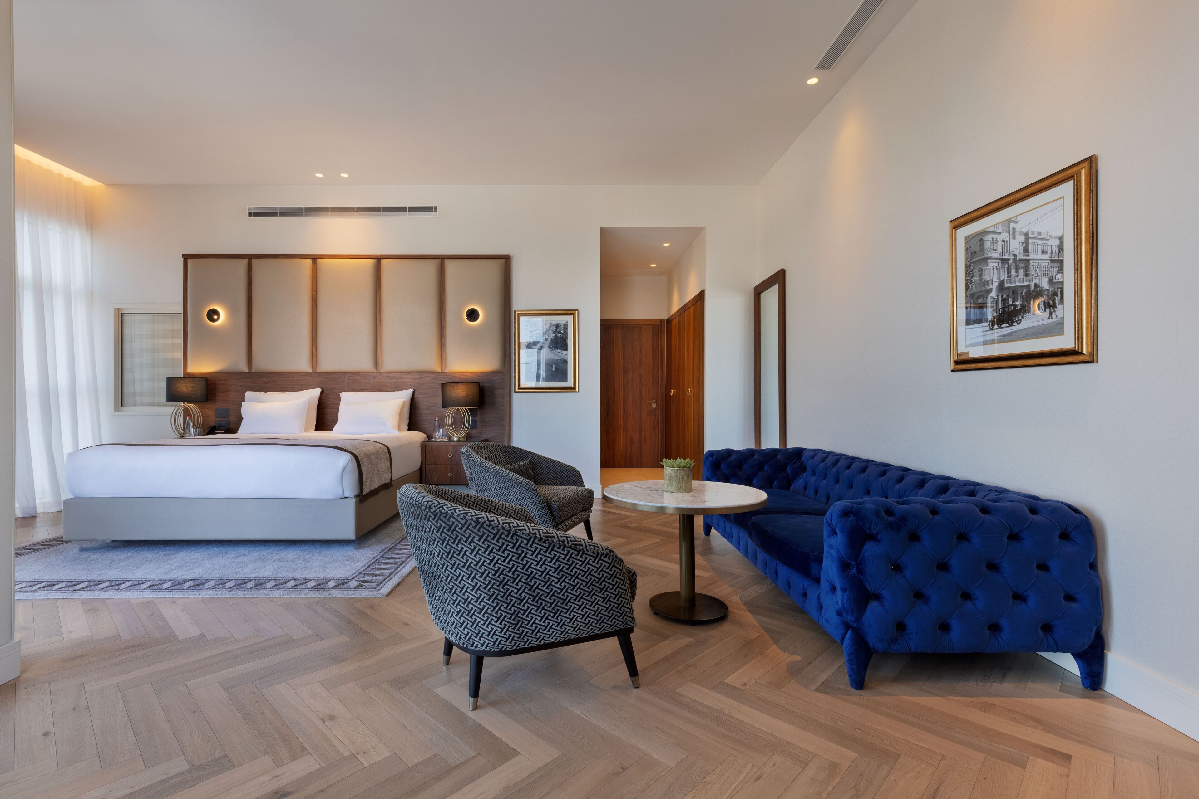 Une chambre d’hôtel moderne avec un grand lit, deux fauteuils, un canapé bleu, une table basse ronde et un tableau encadré au mur.