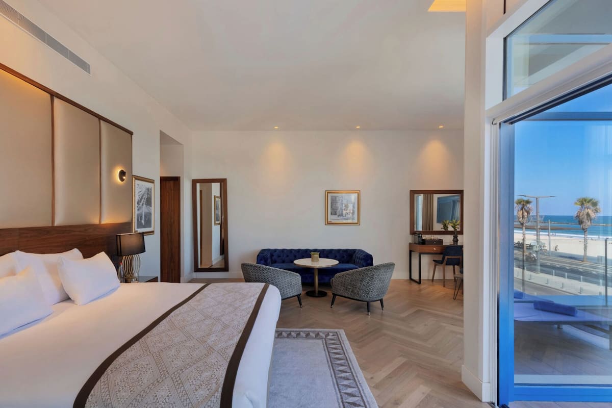 A hotel room with a large bed, a sofa, a small round table, and a desk. There is a large window offering a view of the beach.