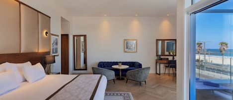 Habitación de hotel con una cama grande, un sofá, una mesita redonda y un escritorio. Una ventana amplia ofrece vistas a la playa.