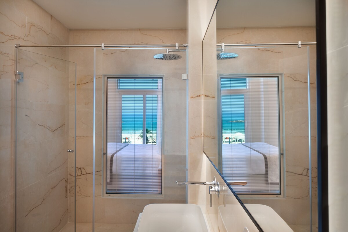 Un baño moderno con cabina de ducha de vidrio, un espejo grande y vistas a la playa a través de la ventana.