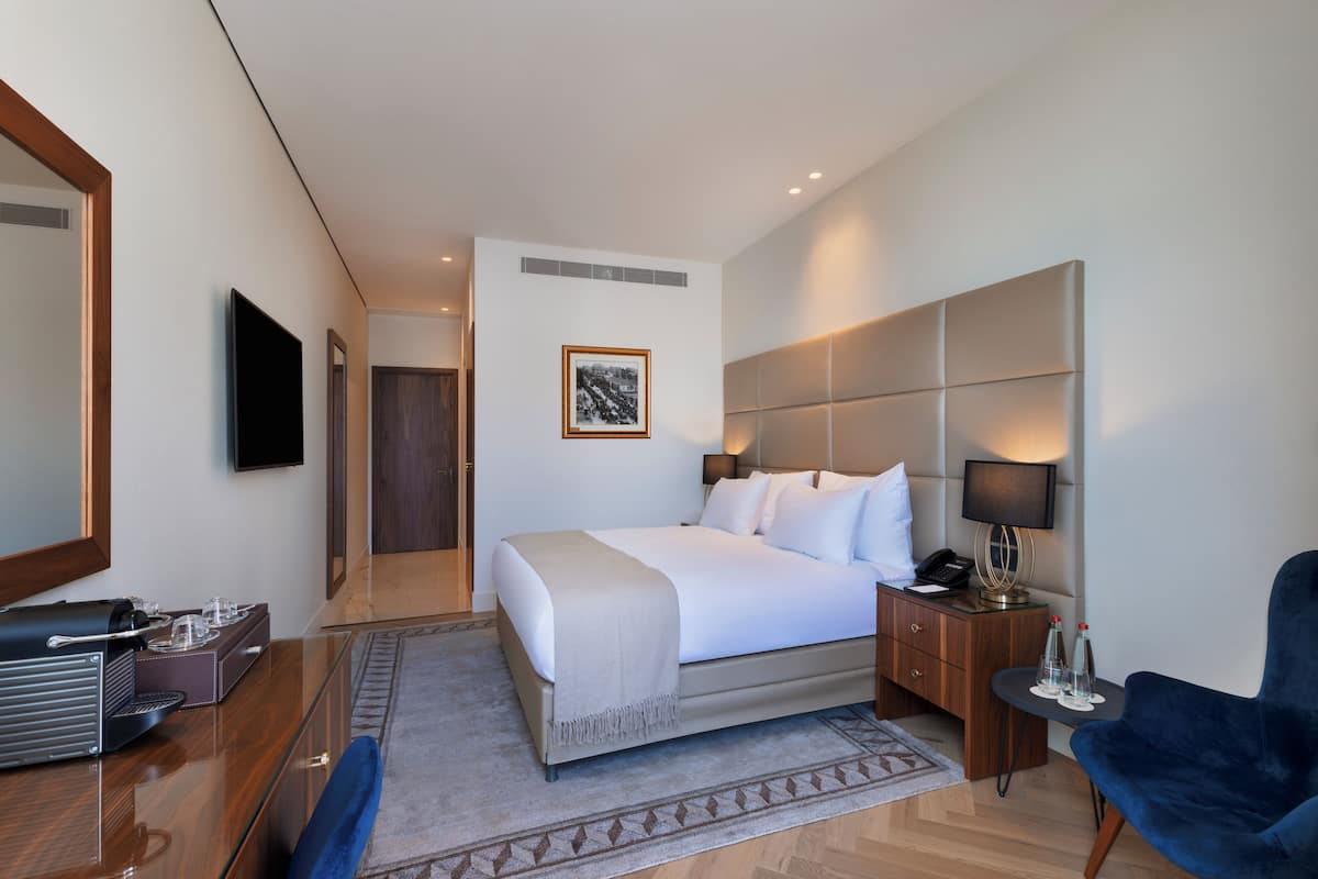 A hotel room with a large bed, bedside tables, a desk with a computer, a chair, and a television.