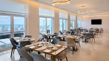 Un vaste espace dédié à la salle à manger, avec de nombreuses tables dressées, de grandes fenêtres offrant une vue sur l’océan et des lustres modernes.
