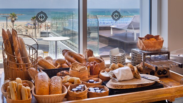 A variety of breads and pastries on a counter with a view of the beach and ocean through large windows. - Herbert Samuel Opera Tel Aviv (Tel Aviv)