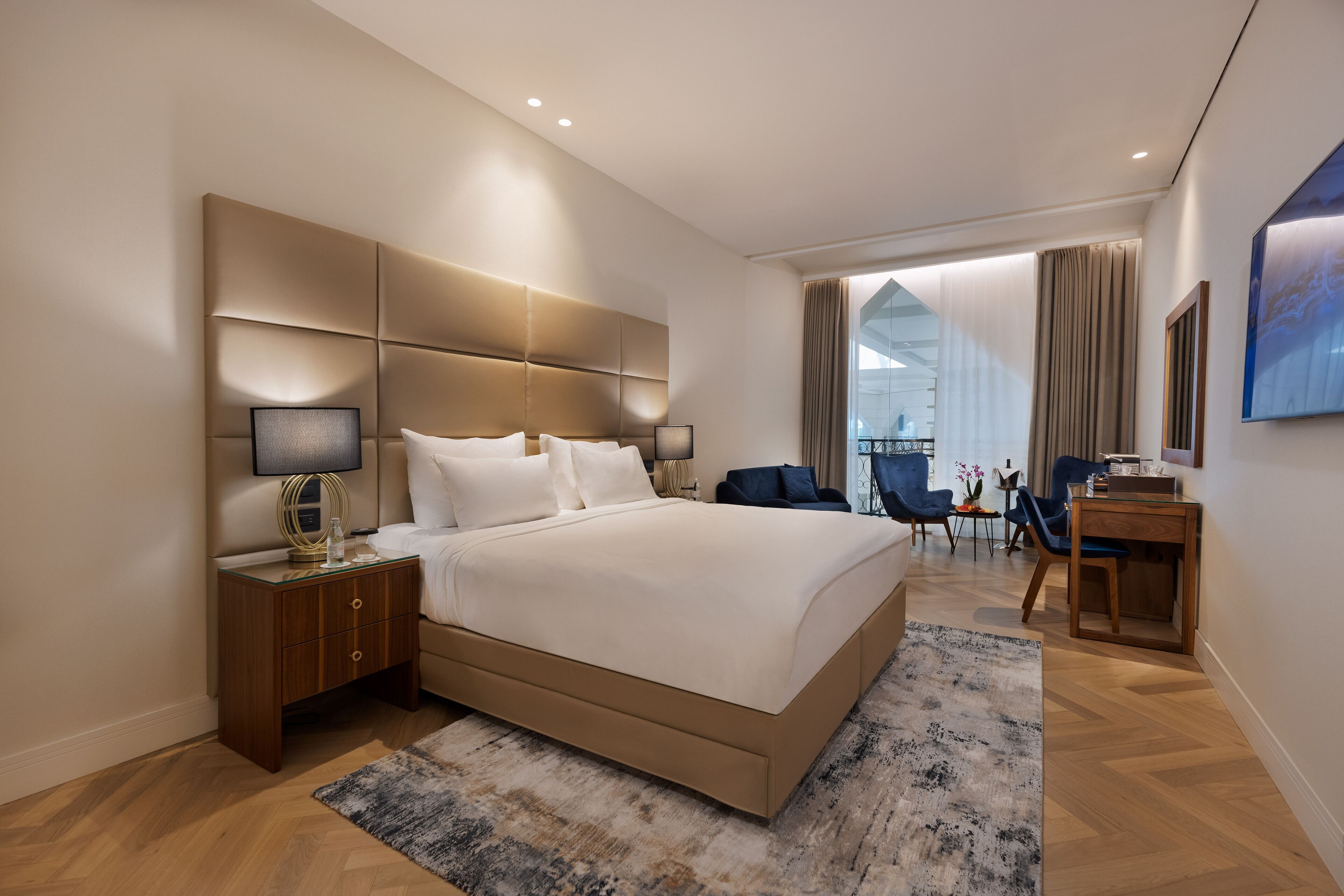 A modern hotel room with a large bed, a nightstand, a desk, and a view of the city through large windows.