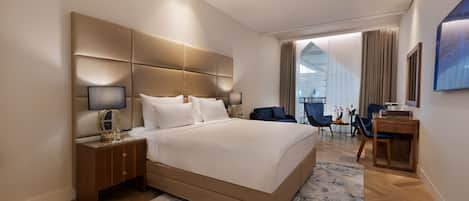 A modern hotel room with a large bed, a nightstand, a desk, and a view of the city through large windows.