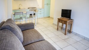 A living room with a sofa, a TV on a wooden stand, and a kitchen in the background. - S102 - Sirolo, new three-room apartment with garden (Sirolo)