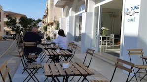 Mesa y sillas en terraza exterior, vista de la calle y un edificio con un cartel que dice "LE DUNE HOTEL CAFE".