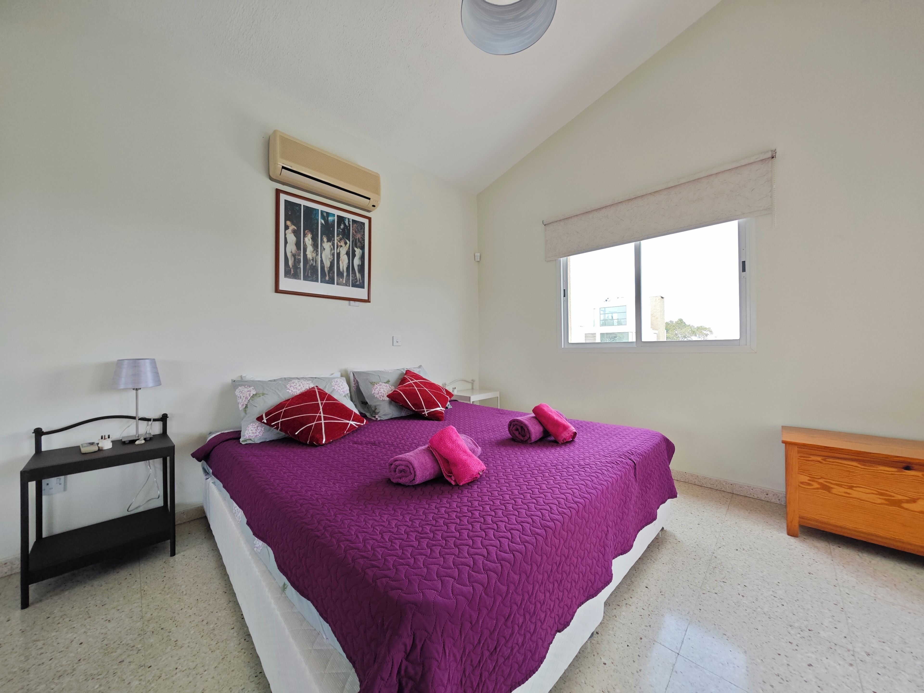 A bedroom with a large bed, a bedside table with a lamp, a window with a view, and an air conditioning unit.