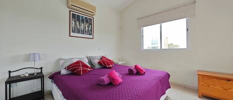 A bedroom with a large bed, a bedside table with a lamp, a window with a view, and an air conditioning unit.