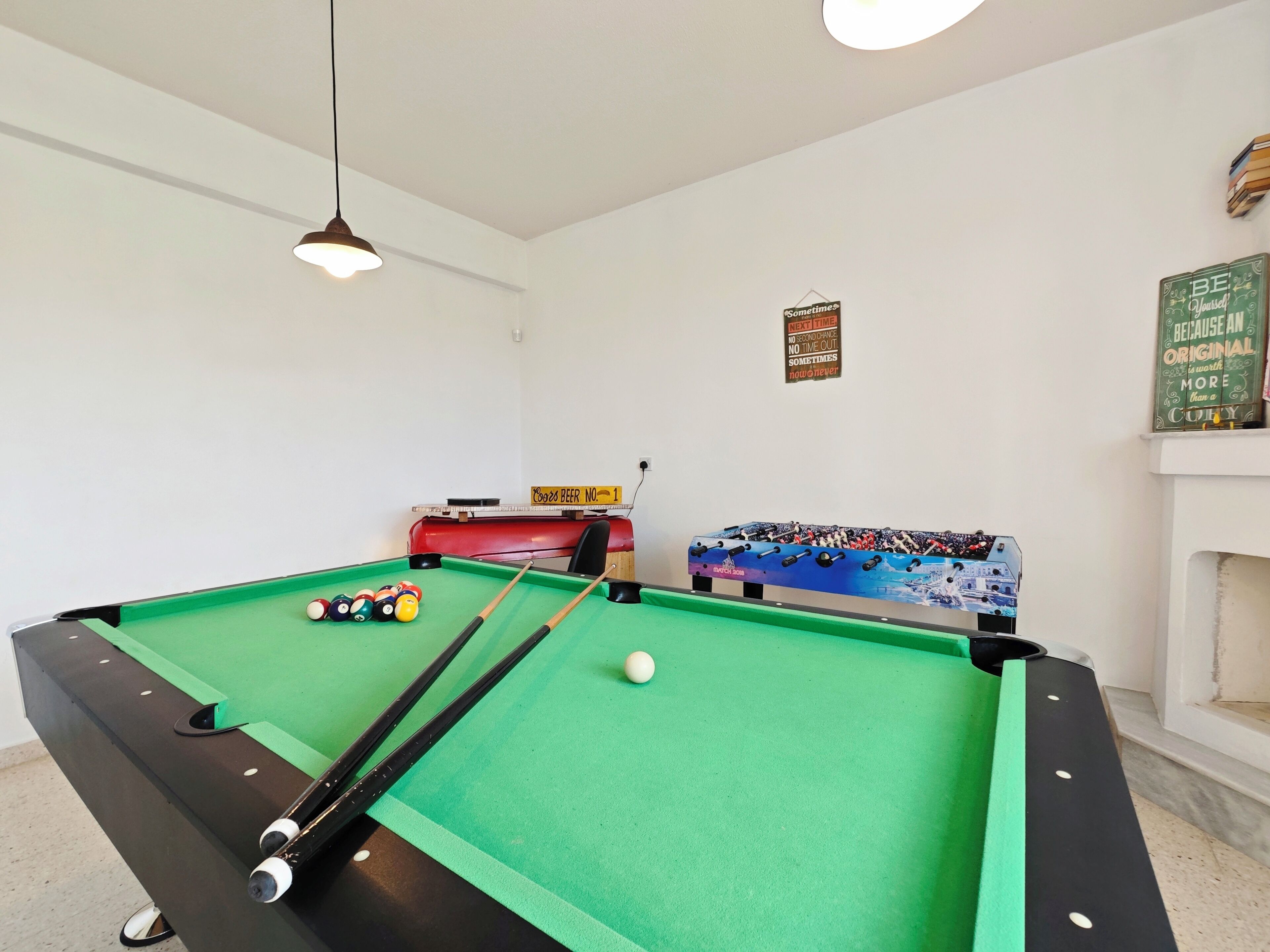 Une table de billard avec des billes, une queue et une machine rouge dans une pièce aux murs blancs et éclairée par un plafonnier.