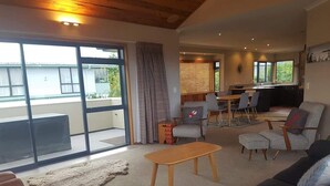 A spacious living room with a large sliding glass door leading to a balcony, a wooden coffee table, a grey sofa, and a dining area with a table and chairs. - View from Fletcher Street (Taupo)
