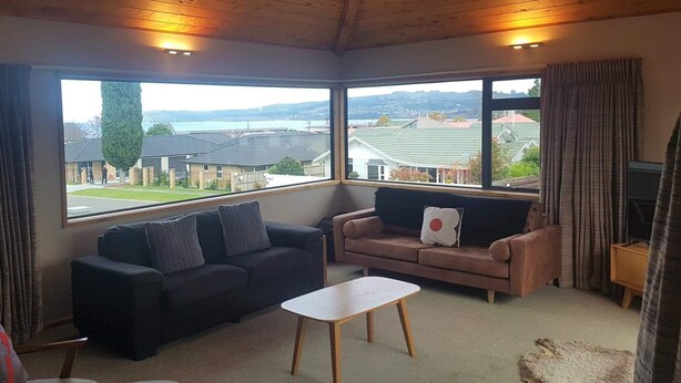 A cozy living room with a wooden ceiling, two sofas, a coffee table, and large windows offering a view of houses and a hill. - View from Fletcher Street (Taupo)
