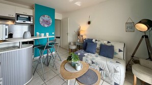 Un espacio habitable compacto con kitchenette, comedor, sofá con cojines geométricos, una pequeña mesa redonda con una planta y una lámpara.