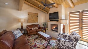 Una sala de estar con un sofá de cuero marrón, un reposapiés estampado, un sillón estampado en marrón y blanco, techo de madera, ventilador de techo, televisión, chimenea y un gran ventanal con persianas.