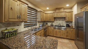 Una cocina con encimeras de granito, armarios de madera y electrodomésticos de acero inoxidable, además de un ventanal con persianas.