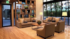A modern lobby with a wooden floor, a sectional sofa, a coffee table, and a bookshelf filled with books and plants. - B Stay Hotel (Bangkok)