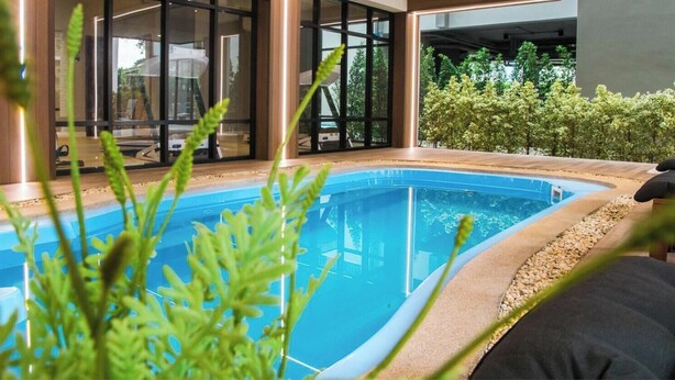 A modern indoor swimming pool with clear glass walls, surrounded by a wooden deck and a small garden area. - B Stay Hotel (Bangkok)