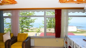 A room with a yellow sofa, a table with chairs, large windows with curtains, and a view of the sea. - Seaside House (Ordu)