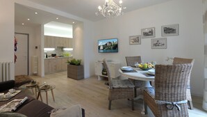 A modern living room with a dining area, a kitchen in the background, and a TV mounted on the wall.