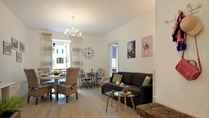 A living room with a dining area, a sofa, a coffee table, and a bench. There are wicker chairs, a chandelier, and a coat rack with hats and a handbag.