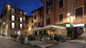 An outdoor cafe area with tables and chairs, surrounded by potted plants and a building with multiple floors and balconies.