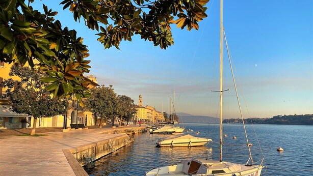 A waterfront promenade with boats docked along the edge, a clear sky, and buildings in the background.