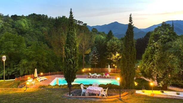 A pool area with lounge chairs and a table, surrounded by trees and mountains in the background. - 4 bed apartment with pool (Pontremoli)