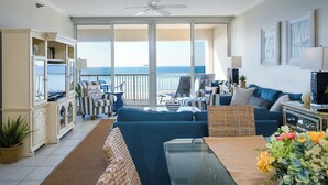 A spacious living room with a large window overlooking the ocean, a ceiling fan, and a dining area with a table and chairs. - Navarre Beach Regency 304 - Eat. Beach. Sleep. Repeat. (Navarre)