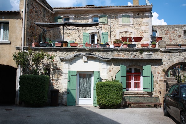 Un bâtiment en pierre doté d’un balcon, de plantes en pot et d’une voiture garée devant.