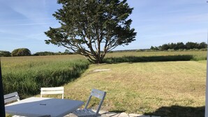 An outdoor patio with a stone floor, white furniture, and a view of a grassy field and a large tree.