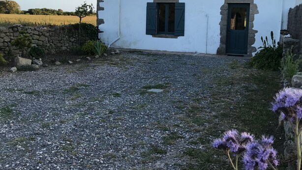 A white building with a stone wall, a gravel driveway, and a field in the background.