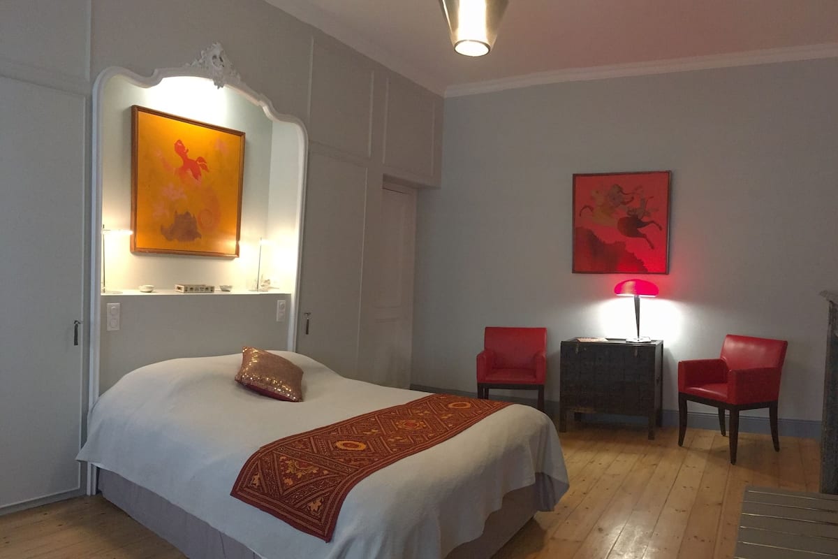 A hotel room with a bed, a red chair, a small table, and artwork on the walls.