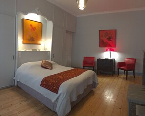 A hotel room with a bed, a red chair, a small table, and artwork on the walls. - Les Chambres d'Hôtes du Palais (Douai)