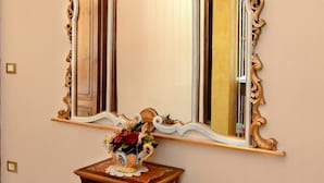 Un espejo con un marco ornamentado fijado a la pared, una pequeña mesa de madera con un jarrón de flores debajo y un suelo a cuadros.