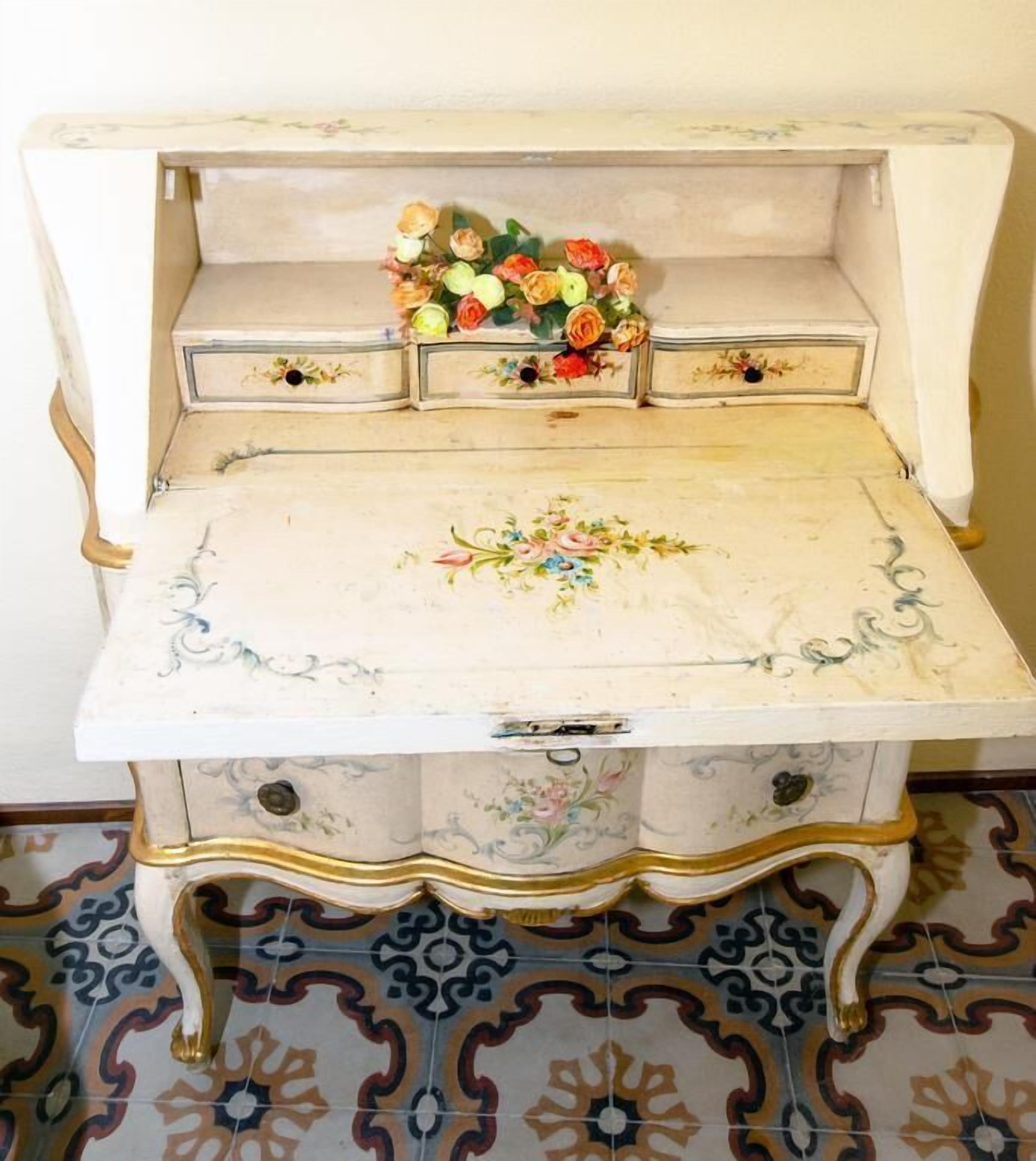 Un bureau de style ancien, orné de motifs floraux, doté de tiroirs et sur lequel est posé un vase rempli de fleurs.