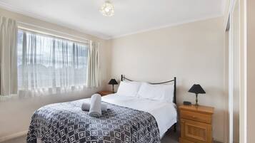 Uma cama bem arrumada com colcha estampada, duas luminárias de cabeceira, uma mesinha de madeira e cortinas leves em uma janela.