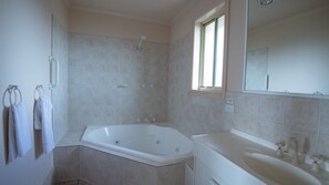 Ein Badezimmer mit einer großen Badewanne, einer Dusche und einem Waschbecken. Handtücher hängen an einem Ablagekorb und ein Fenster lässt natürliches Licht herein.