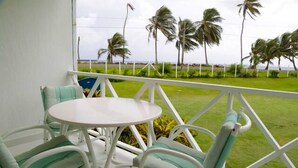 Un balcón con mesa y sillas blancas, con vistas a una zona verde con palmeras y al océano.