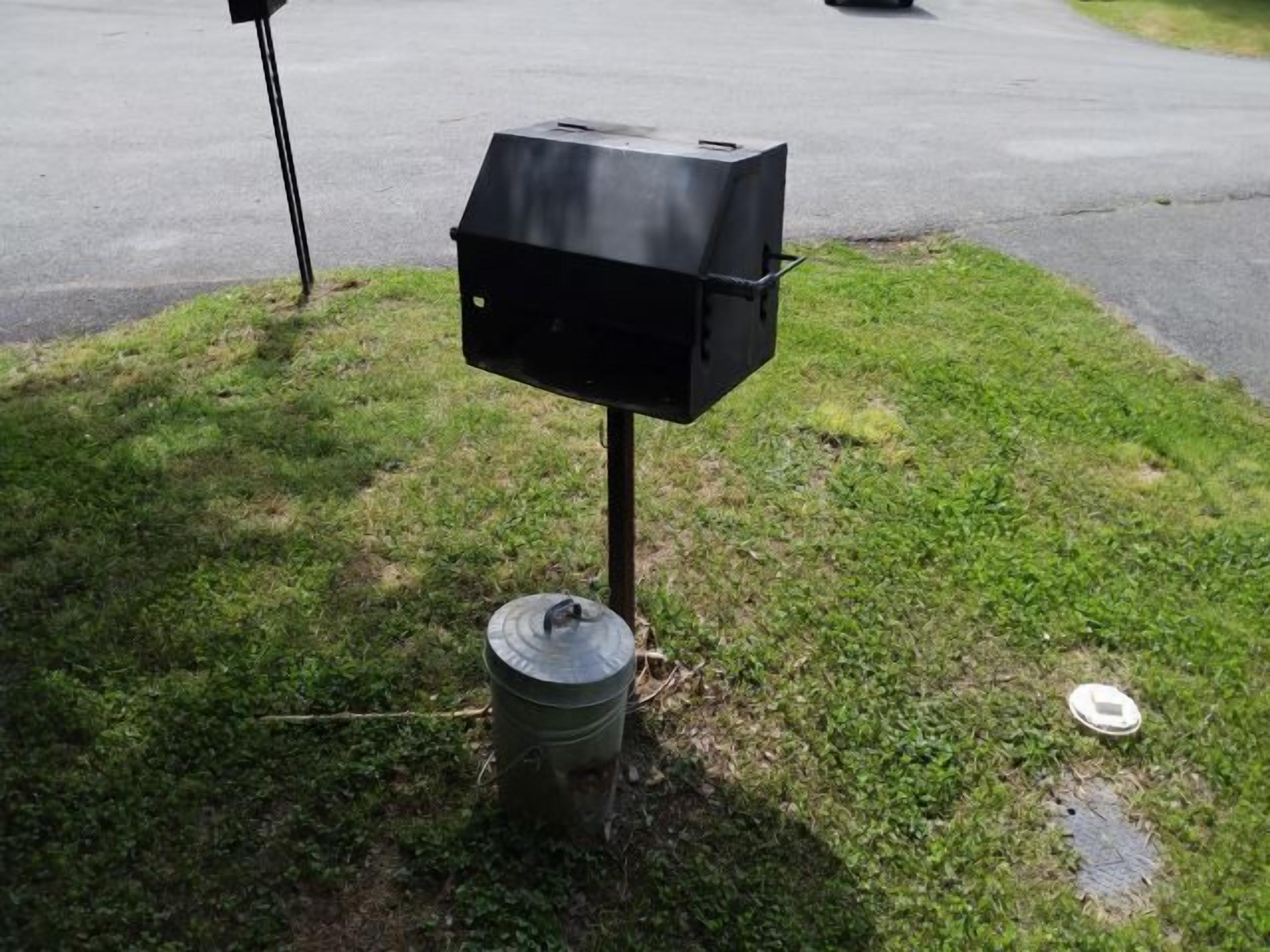 Una parrilla negra al aire libre sobre una zona cubierta de césped, con un bote de basura metálico cerca.