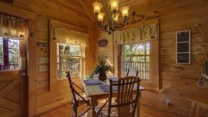 Interior de una cabaña de madera con una mesa de comedor, sillas, una araña y ventanas con cortinas.