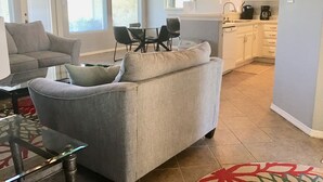 Un salon avec un canapé gris et une table basse en verre, un coin repas avec une table et des chaises, et une cuisine avec des meubles blancs et des appareils électroménagers.