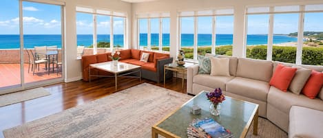 A spacious living room with a large window overlooking the ocean, a sofa, a coffee table, and a dining area with chairs.