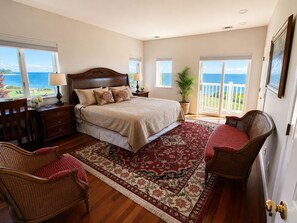 A bedroom with a large bed, a sitting area with a chair, a dresser, and a view of the ocean.