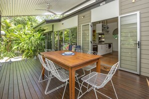 A wooden deck with a dining table and chairs, a kitchen area, and a glass door leading to a garden.