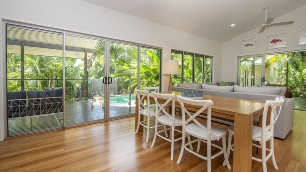 A spacious living area with a wooden floor, a dining table with chairs, a sofa, and a ceiling fan. Large windows provide a view of lush greenery outside.