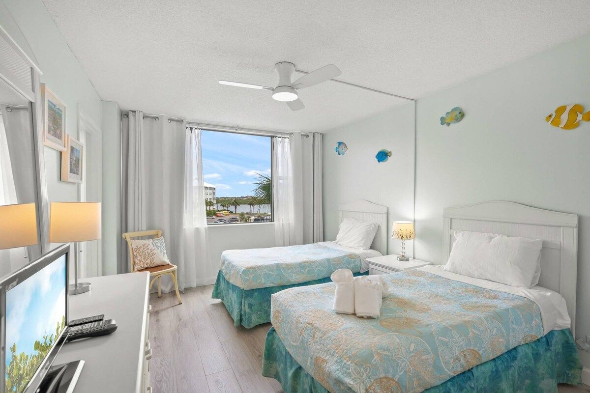 A hotel room with two beds, a desk with a computer, a chair, a window with curtains, and a ceiling fan.
