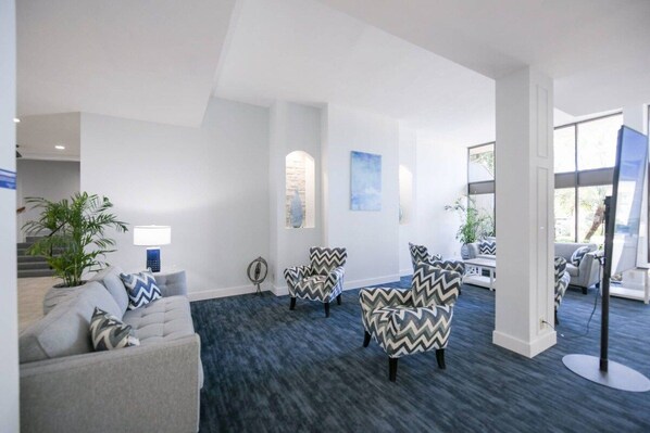 A modern hotel lobby with a grey sofa, patterned armchairs, a potted plant, and a large window.