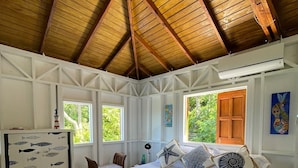 A bedroom with a wooden ceiling, a bed with a blue and white fish-patterned bedspread, two windows, and a wall-mounted air conditioner. - Las Palmas in Kitten's Warf (Providencia)