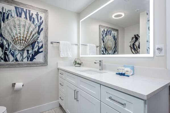 A modern bathroom with a large mirror, white cabinetry, and a framed seashore painting.
