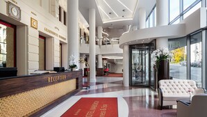 A Grand Hotel lobby with a reception desk, a red carpet with the hotel name, a white sofa, and a large window offering a view of the street. - Hotel Nabokov (Marianske Lazne)